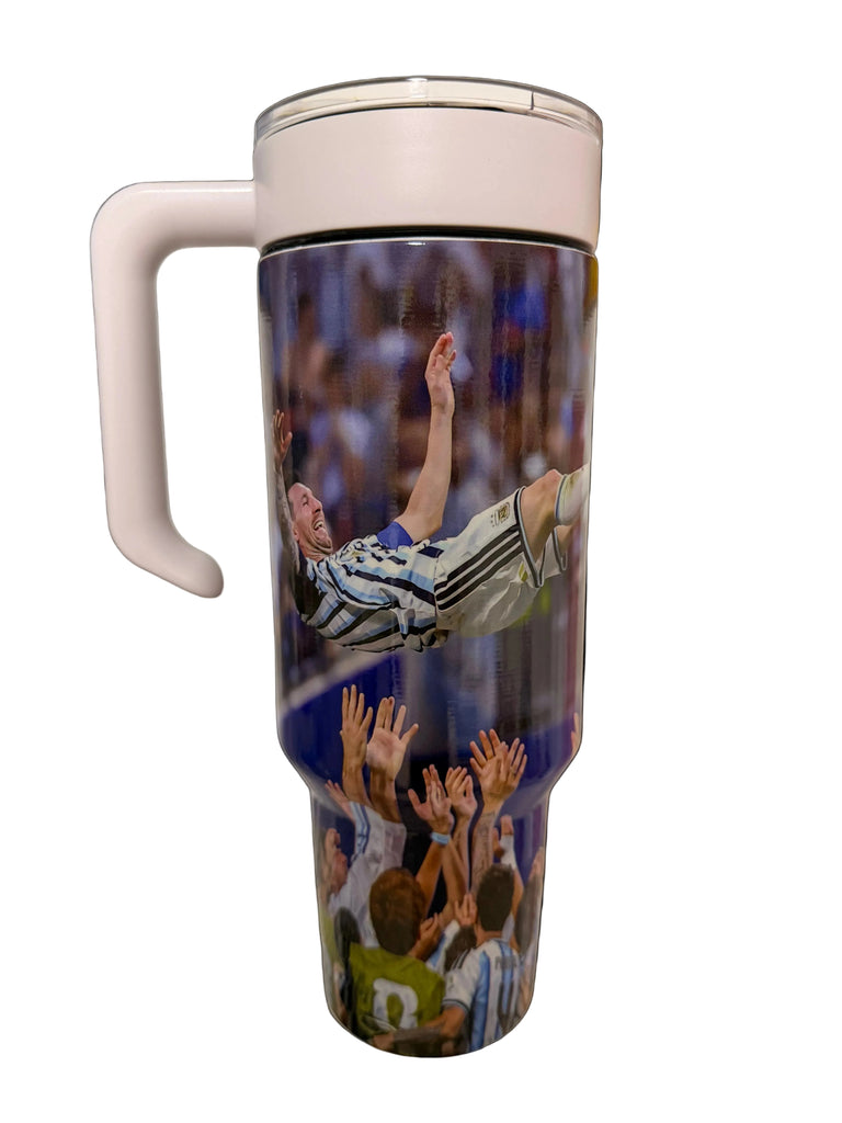 A handled travel tumbler with a printed soccer player design, shown lying on a white shelf against a white wall.